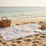 Beach scene with a colorful towel, picnic basket, and hat on sand near ocean.