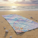 Pastel mermaid tail beach towel laid on sand at sunset with scattered shells