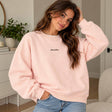 Woman in a pink crew neck sweatshirt with a dainty Aube de Paris script, hands in her jeans pockets