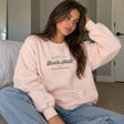 Woman in a pink crew neck sweatshirt with a vintage script chest print, sitting in a bedroom