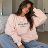Woman in a pink crew neck sweatshirt with a vintage script chest print, sitting in a bedroom