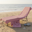 A pink beach towel draped over a lounge chair on a sandy beach, with matching pink tote bags and seashells nearby and the ocean in the background.