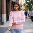 Woman in a pink brushed wool crew neck sweater and light jeans smiling and tugging her sleeve on a sidewalk