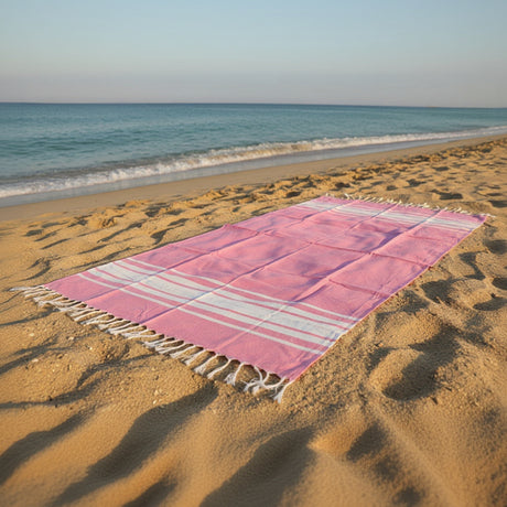 Soft pink striped beach towel with tassel edges on sandy beach with ocean waves