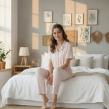 Woman in a light pink cotton pyjama set standing in a bright bedroom with plants and decor
