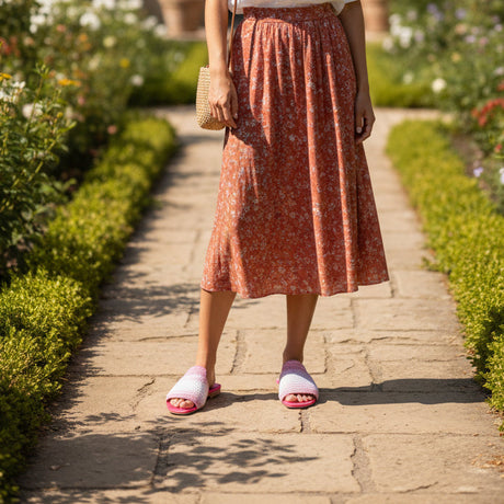 Person wearing a rust-colored floral skirt and pink handmade slippers walking on a paved path in a garden