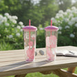 Two clear heart print tumblers with pink lids and straws on a garden table