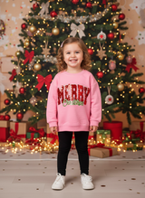 Girl in a pink Merry Christmas sweatshirt set smiling in front of a red and gold Christmas tree