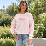 Woman in a pale pink sweatshirt with pink sequin lettering, standing in a sunny garden