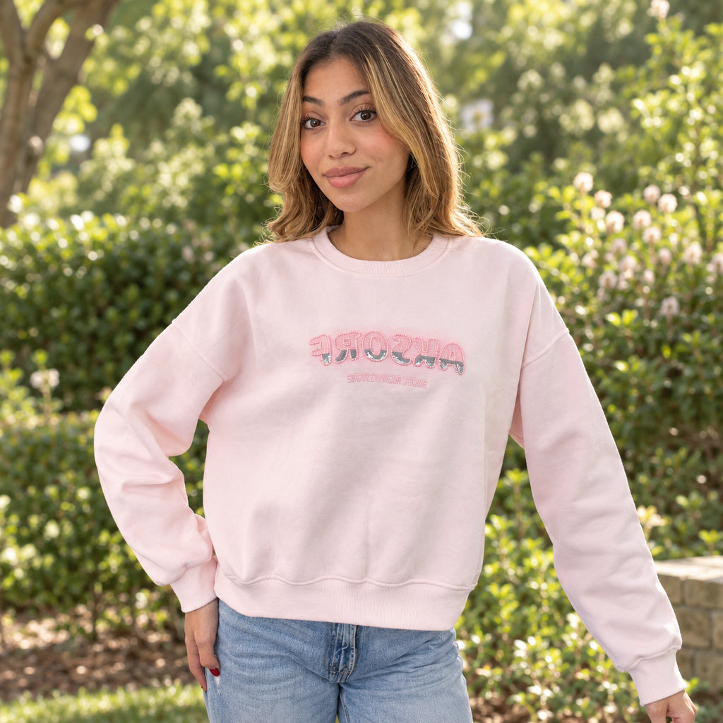 Pale pink crew neck sweatshirt with pink sequin letters, worn with light jeans outdoors