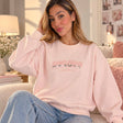 Woman sitting on a sofa in a pale pink sweatshirt with a silver sequin slogan