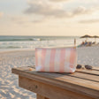 A pink and white striped zippered pouch bag sits on a wooden beach table at the beach