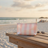 A pink and white striped zippered pouch bag sits on a wooden beach table at the beach