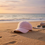 Soft pink cotton summer cap on sand beside a towel and sunglasses