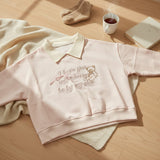 Woman wearing a pink collared sweatshirt with an embroidered teddy bear and script slogan