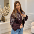 Woman in an open plum bomber jacket with white beaded flowers over a beige crop top and jeans