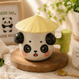 Ceramic mug with panda design and straw hat on a wooden coaster, surrounded by decorative items.