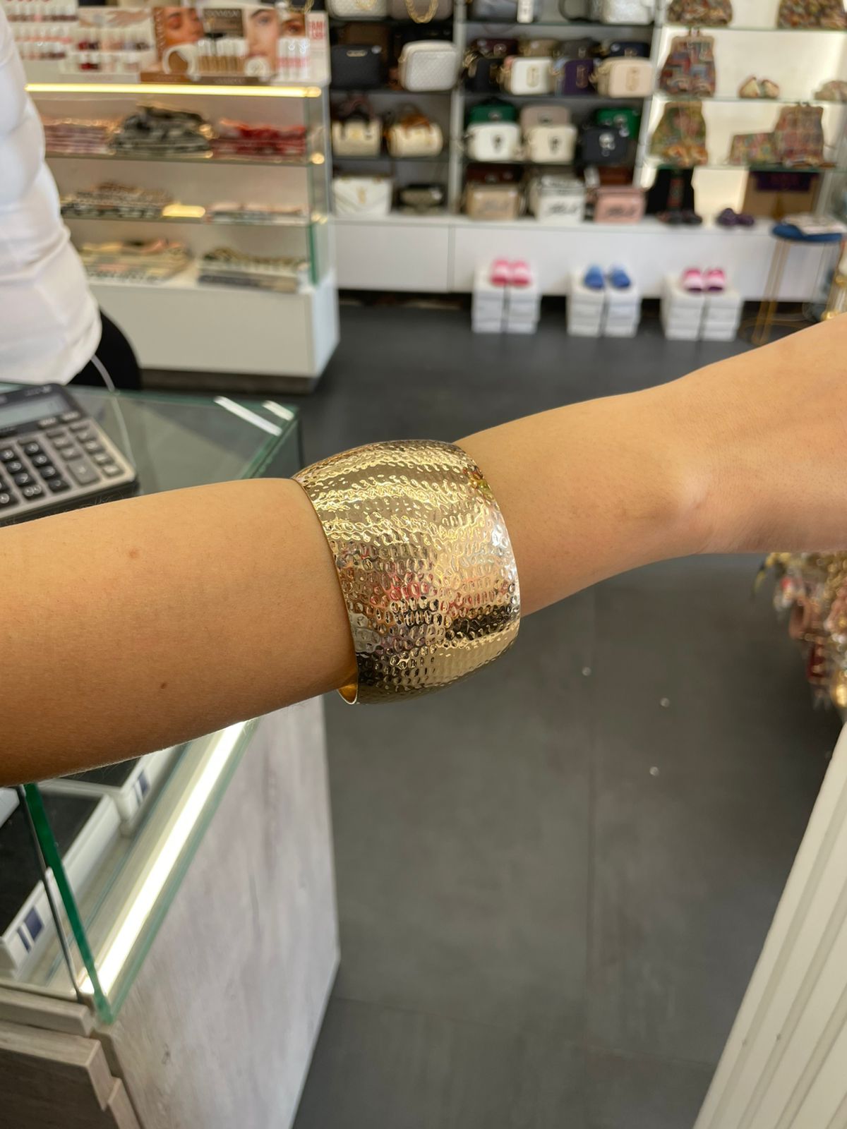 Gold polished wide cuff bracelet on a wrist with a store interior in the background