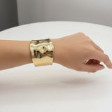 Gold polished wide cuff bracelet on a wrist against a neutral background
