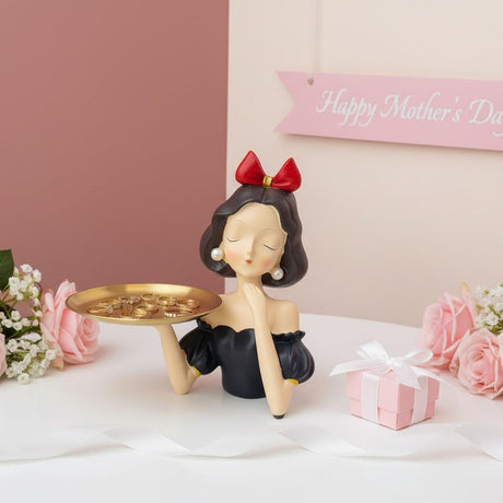 Black bobbed figure with a red bow, gold rings laid across her tray among roses