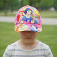 Pink kids baseball cap printed with three cartoon fairytale princesses, worn by a child in a park