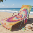 A metal beach lounge chair on a sandy beach with the ocean in the background. The chair is covered in a bright tie-dye blanket featuring a spiral pattern of pink, yellow, orange, green, and purple.