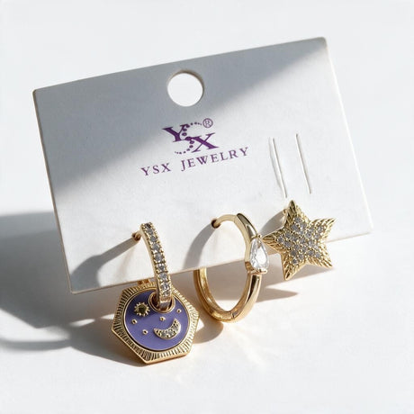 Three gold single earrings on a card with a purple moon charm drop, a crystal teardrop hoop and a pave star stud