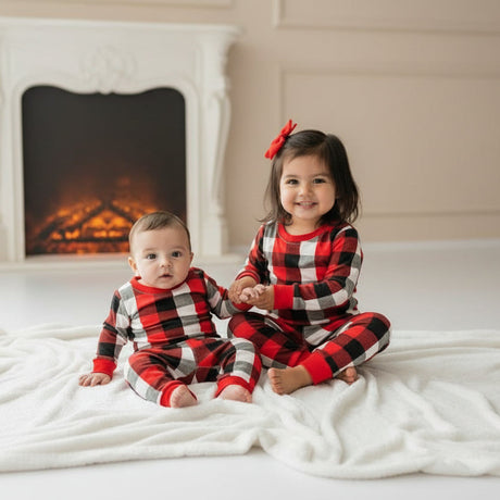 Baby and toddler sister in matching red check Christmas pyjamas by a white fireplace