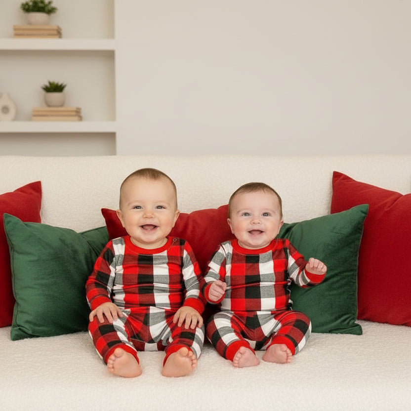 Two babies sitting side by side in red and black buffalo check pyjamas on a sofa