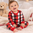 Smiling baby in red and black checkered pyjamas sitting on a cozy bed with a teddy bear