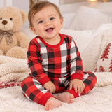 Smiling baby in red and black checkered pyjamas sitting on a cozy bed with a teddy bear