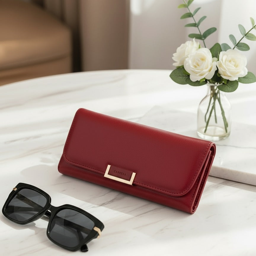 Red wallet with gold accents and black sunglasses on a white surface, next to a vase of white flowers