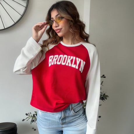 Woman wearing a red and white raglan Brooklyn sweatshirt with light wash jeans and tinted glasses