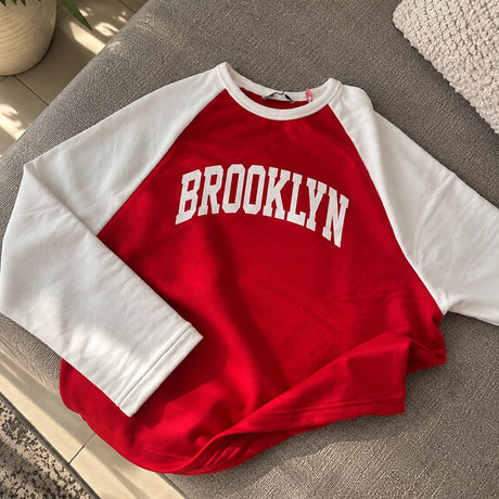 Red Brooklyn sweatshirt with white raglan sleeves laid flat on a grey sofa
