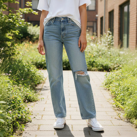 Person wearing light blue ripped jeans with a slight tear on a paved path with greenery