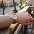 A wristwatch with a rose gold-toned case and a two-tone silver and rose gold metal link bracelet, worn on a person's wrist in an outdoor cafe setting with a blurred street background.