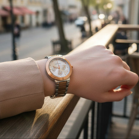 A wristwatch with a rose gold-toned case and a two-tone silver and rose gold metal link bracelet, worn on a person's wrist in an outdoor cafe setting with a blurred street background.