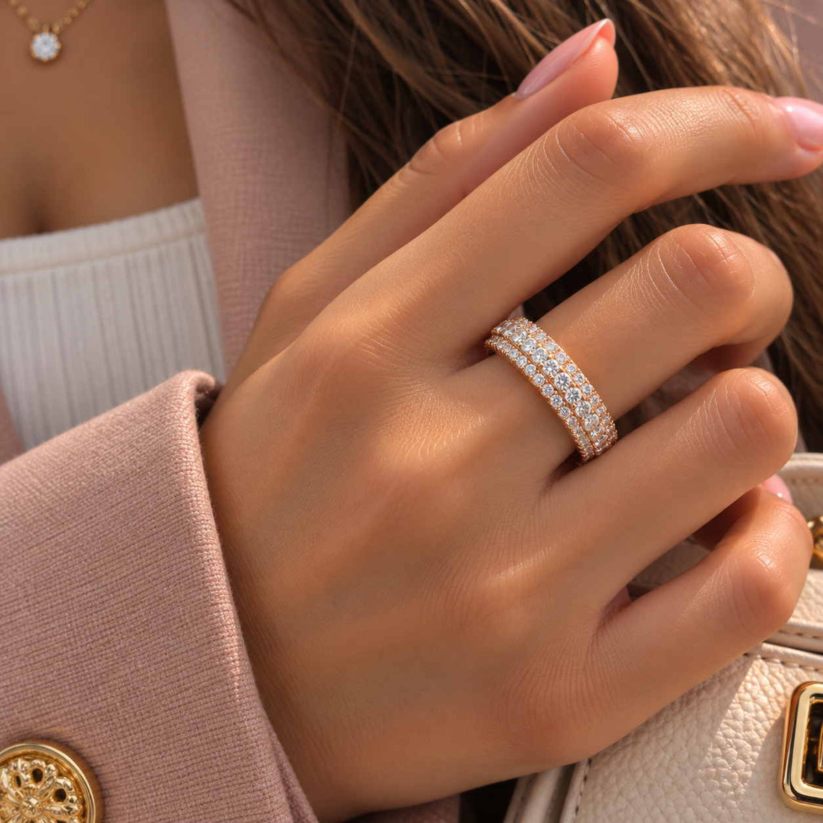Hand wearing a rose gold band ring with three rows of round crystals, resting on a pink blazer