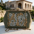 Sage green kiss lock clutch with Arabic calligraphy embroidery in front of an arched stone house