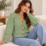 Sage green pointelle cardigan with tortoiseshell buttons, worn sitting on a cream sofa