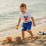 Child playing on a beach with sandcastle materials, wearing a 'Sea Friends' shirt and blue shorts.