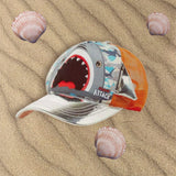 Shark face cap with a white brim and orange mesh back resting on sand beside seashells