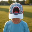 Boy wearing a grey shark face baseball cap with blue camo panels and an orange mesh back