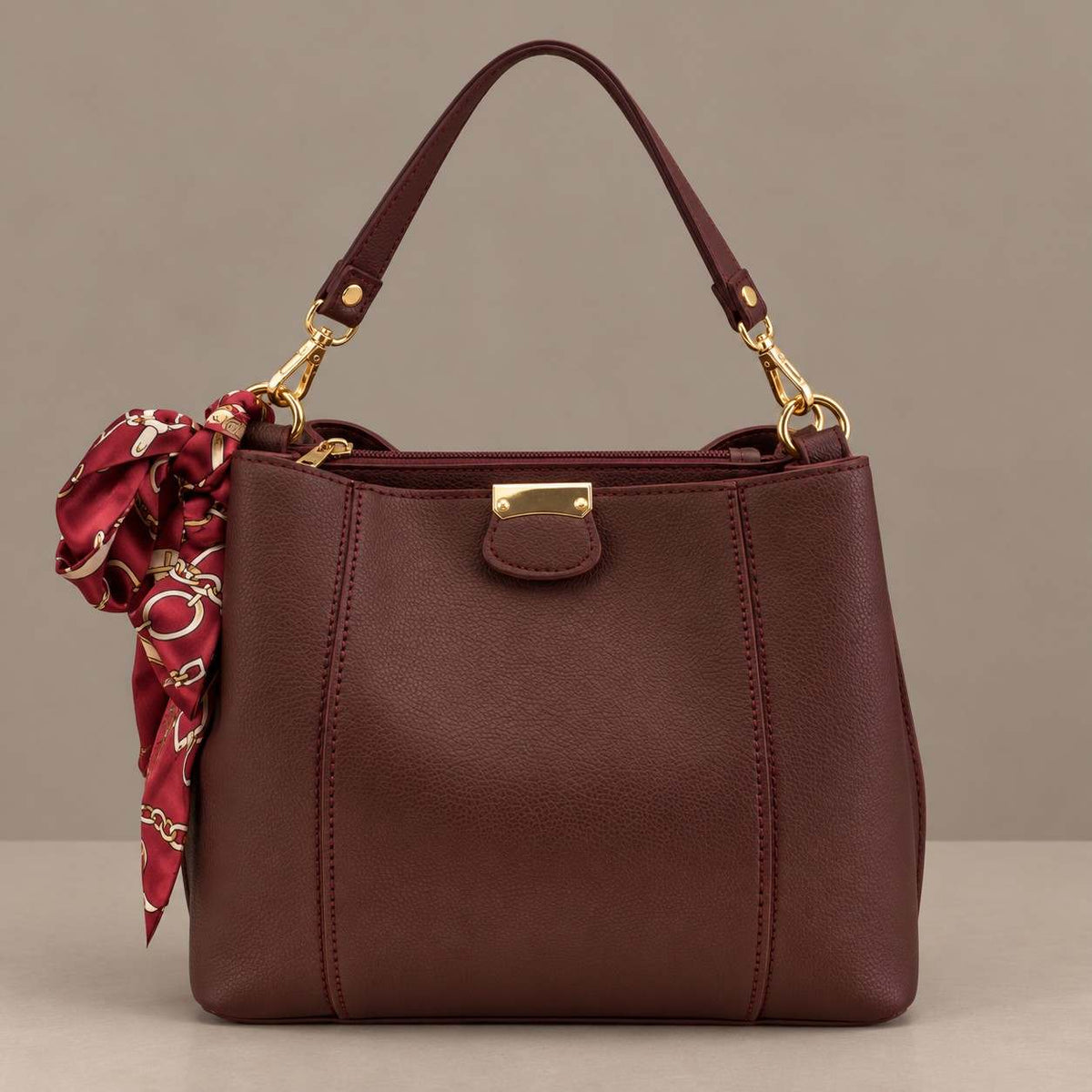 Burgundy top handle bag with gold clips and a printed scarf tied at the side