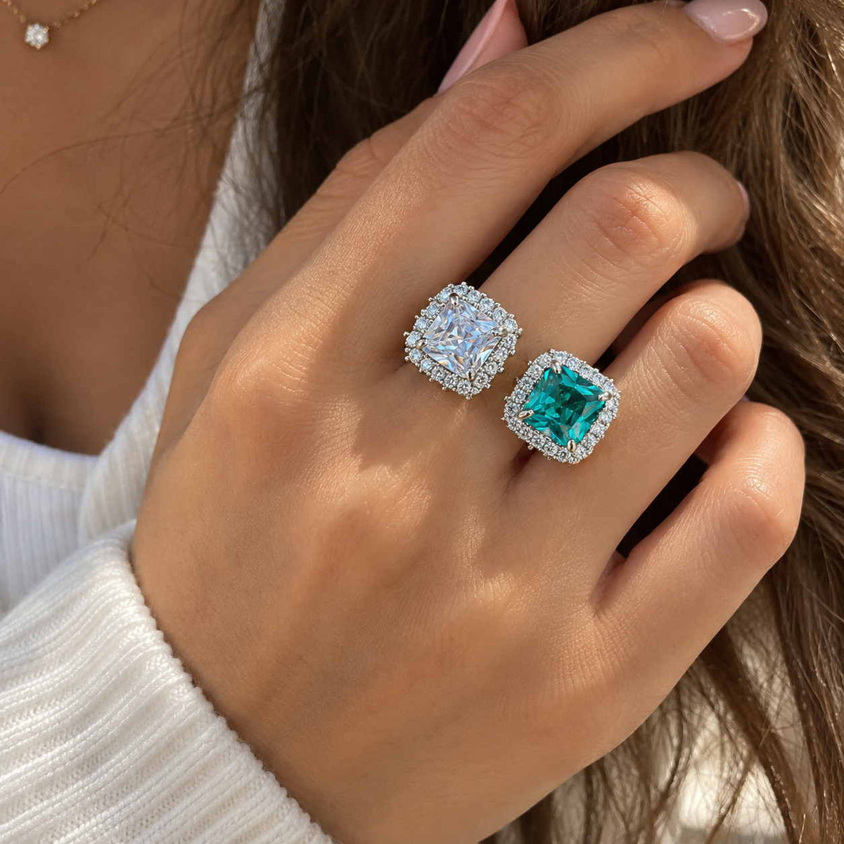 Hand wearing a silver ring with a clear square crystal and a teal stone, each framed by small crystals