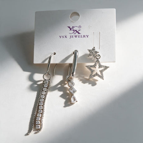 Three silver single earrings on a card with a crystal chain drop, a charm huggie and an open star stud