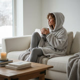 Silver grey sherpa hooded blanket worn seated on a sofa by a bright winter window
