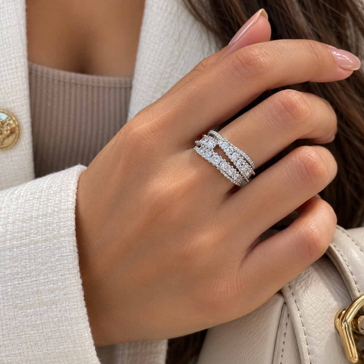 Hand wearing a silver crossover ring with layered rows of round and square crystals, holding a cream bag