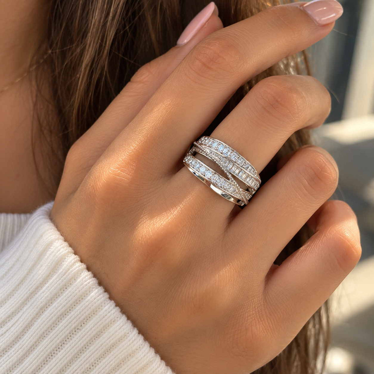 Hand wearing a wide silver ring with sweeping rows of round and baguette crystals, over a white knit sleeve