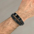 Black leather bracelet with metal clasp on a wrist against a grey background
